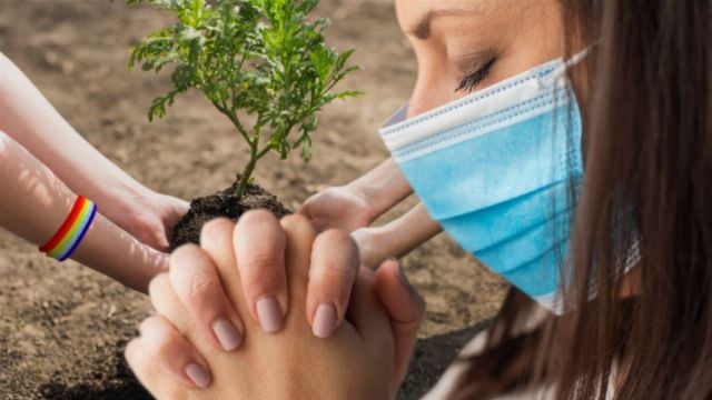 **** Persono portanta medicinan maskon videblas kun fermitaj okuloj kaj kunpremitaj manoj en preĝo. En la fono, manoj de pluraj individuoj videblas plantantaj malgrandan verdan arbon en malhela grundo, simbolante esperon kaj komunumajn klopodojn por restarigi la naturon.