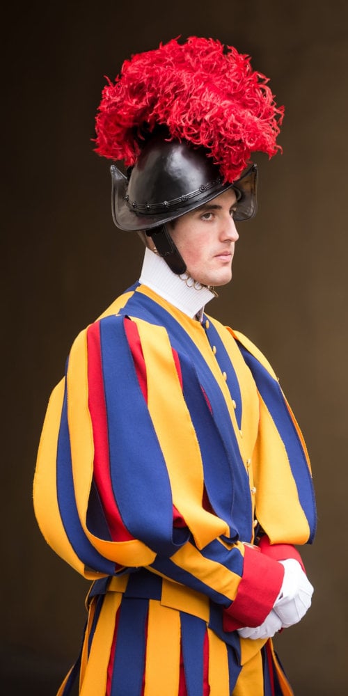 A man in a ceremonial uniform stands in profile. He wears a striking helmet with a voluminous red plume and a multicolored uniform in bold stripes of blue, orange, and yellow. His expression is serious, and he is posed against a neutral brown background.