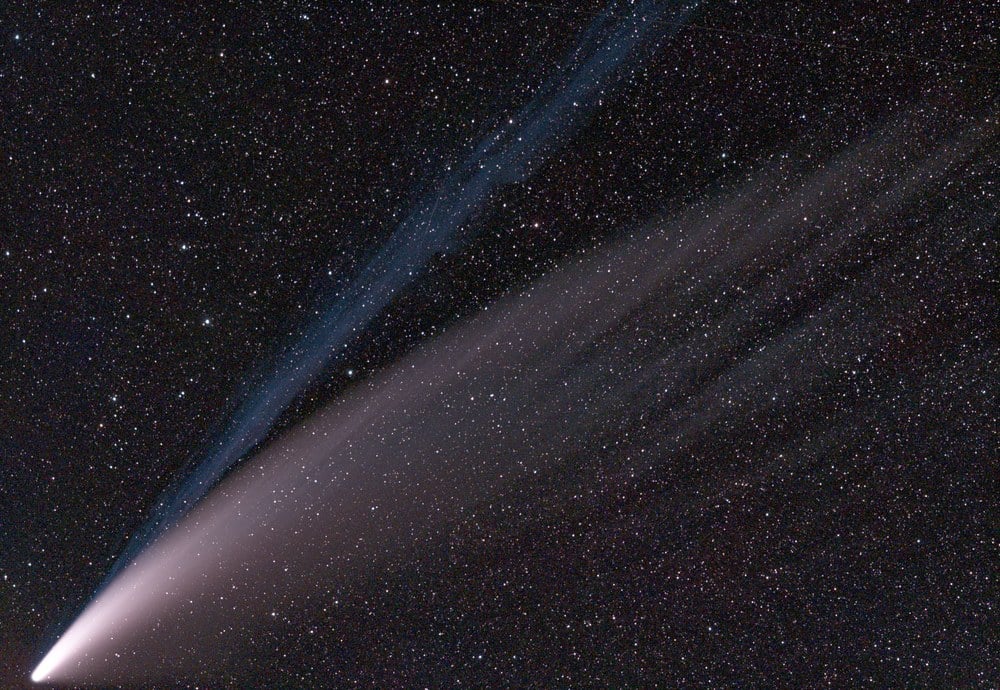 Un cometa con un n&uacute;cleo brillante que emite una cola larga y tenue, con un tel&oacute;n de fondo de innumerables estrellas en un cielo nocturno oscuro.