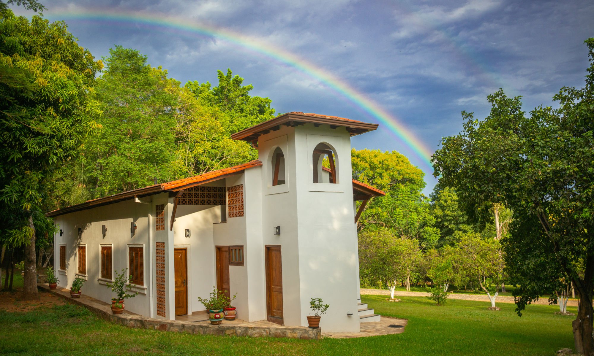 Ein malerisches weißes Stuckhaus mit Terrakotta-Ziegeldach und Holzakzenten, umgeben von üppigem Grün unter einem strahlenden Himmel, über dem sich ein leuchtender doppelter Regenbogen wölbt.